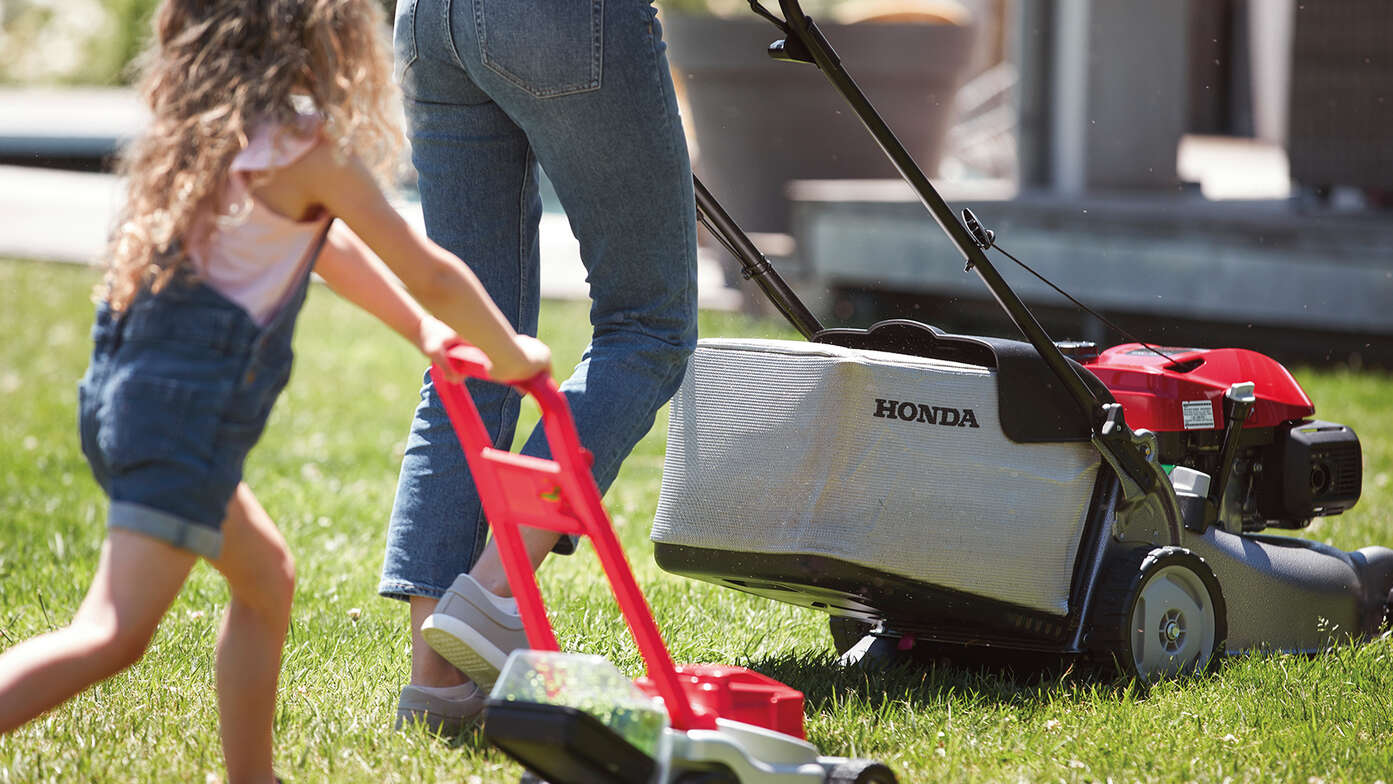 Honda IZY-gräsklippare sedd från sidan med kvinna och barn i trädgårdsmiljö