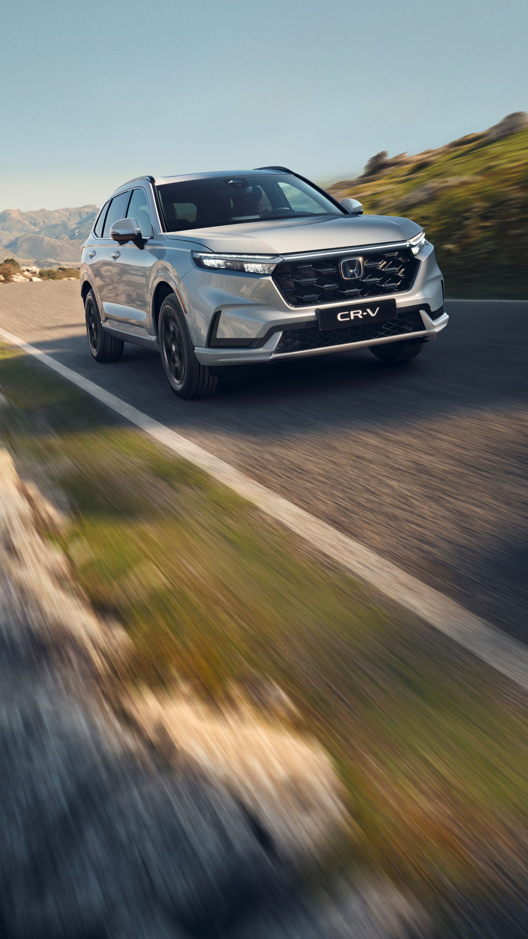 En silvrig Honda CR-V SUV kör på en slingrande bergsväg med karg terräng och berg i bakgrunden. Bilen är i rörelse, vilket skapar en känsla av fart och äventyr i det karga landskapet.
