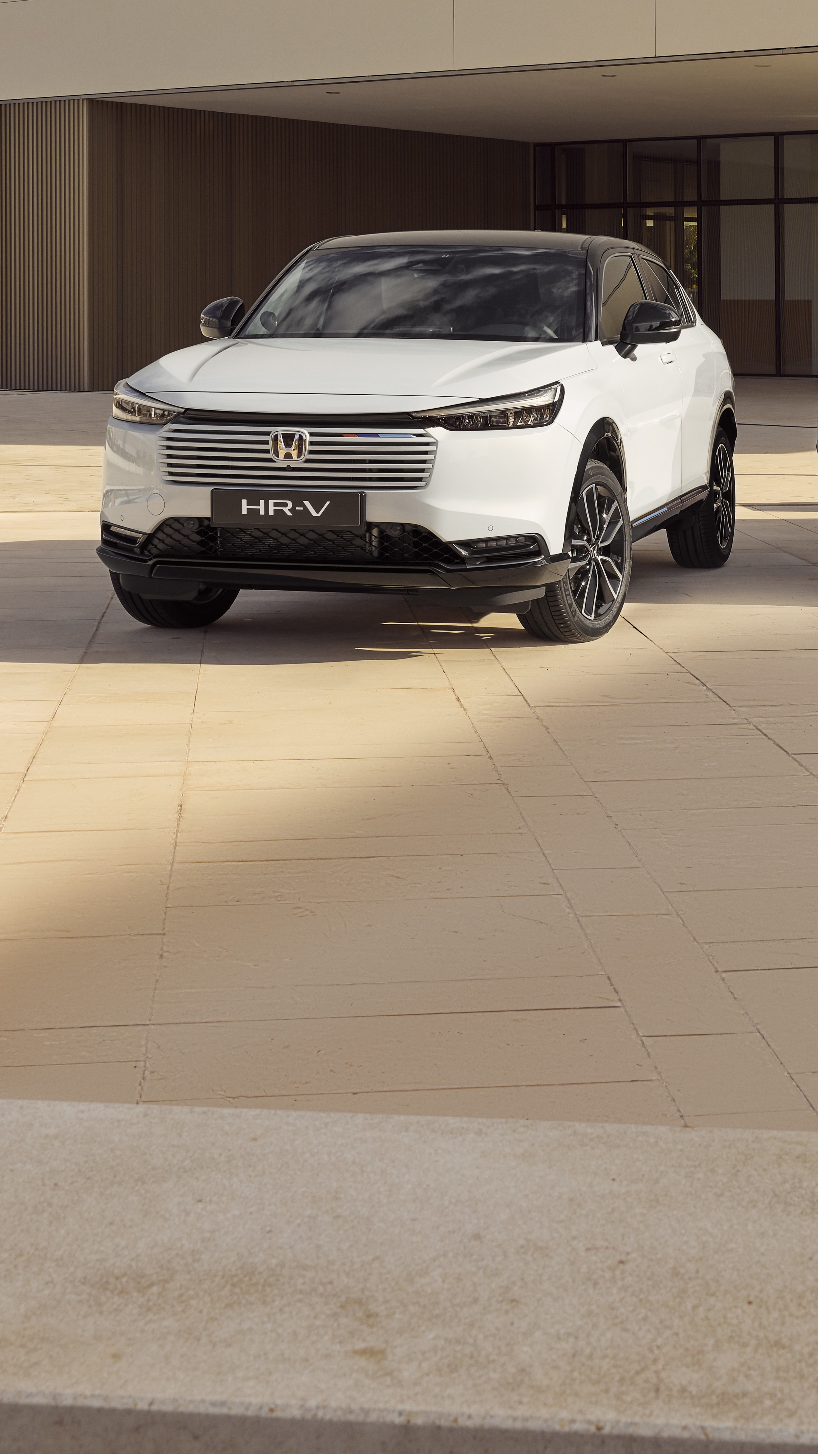 En vit Honda HR-V SUV parkerad framför en modern byggnad med beigekaklade väggar. Bilen står i vinkel för att visa upp sin eleganta design och karakteristiska grill.