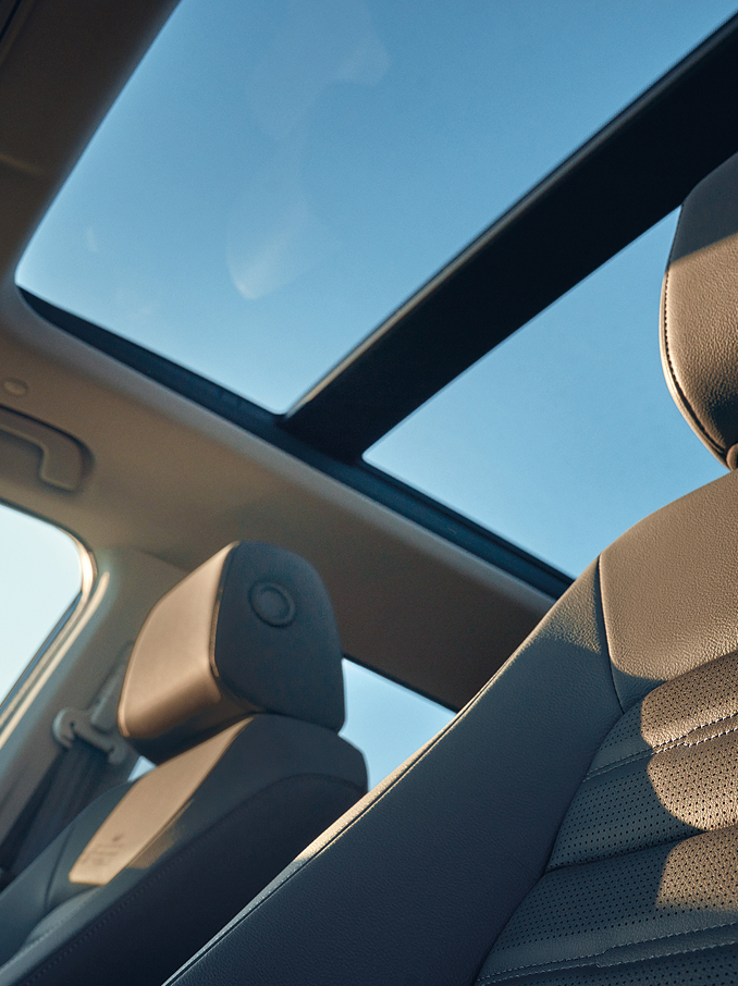 Interiörvy av en bil som visar ett panoramaglastak som släpper in ljus från den blå himlen och solen, med framsäten i läder och nackstöd belysta av naturligt ljus.