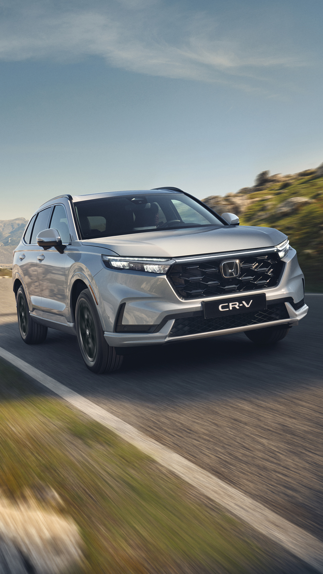 En silverfärgad Honda CR-V SUV kör på en slingrande bergsväg med stenig terräng och berg i bakgrunden under en klarblå himmel.