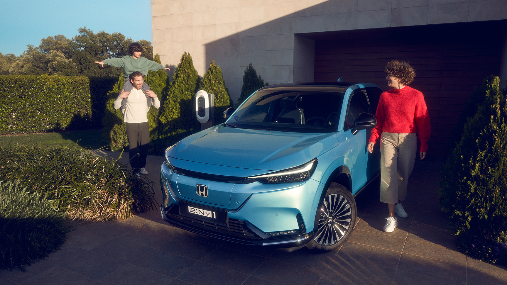 Familj som står bredvid en framåtvänd Honda e:Ny1 parkerad utanför ett hus.