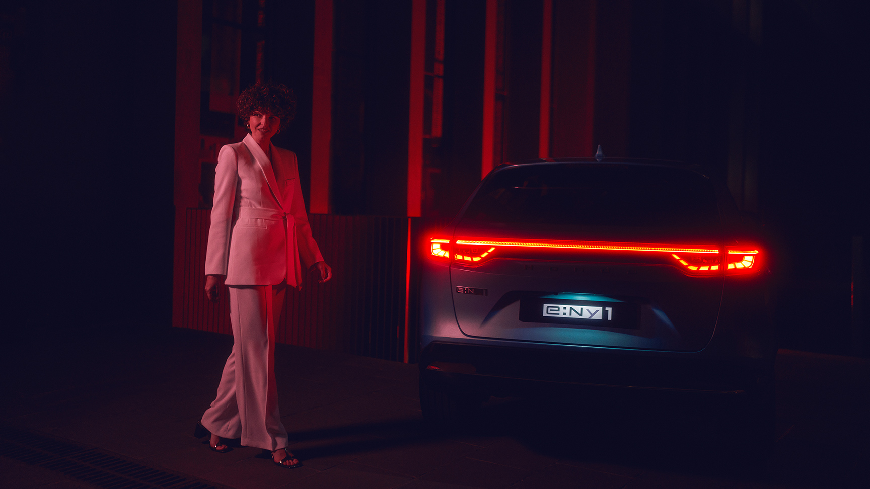 Modell som står bredvid den upplysta baksidan av Honda e:Ny1 på natten.