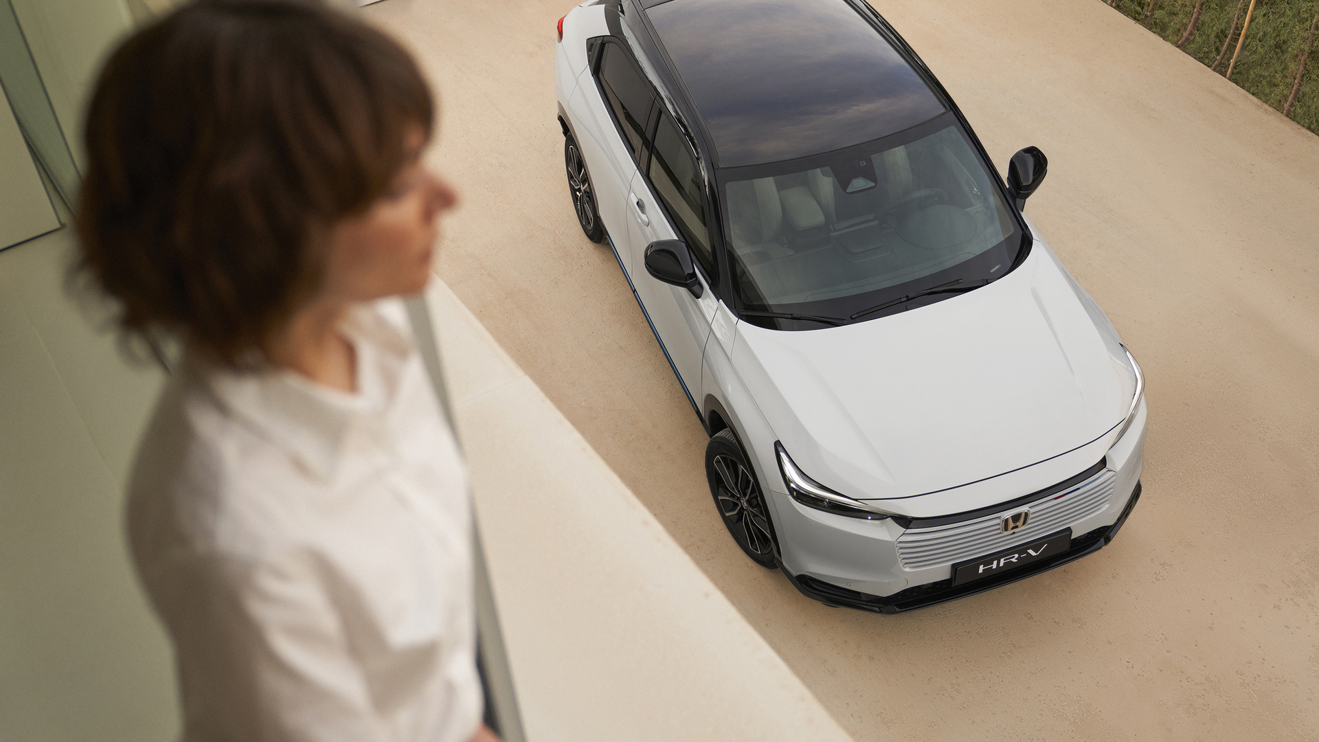 En ovanifrånvy av en vit Honda HR-V med svart tak parkerad på en ljus uppfart, medan en suddig person står inomhus i förgrunden och tittar ner mot bilen.