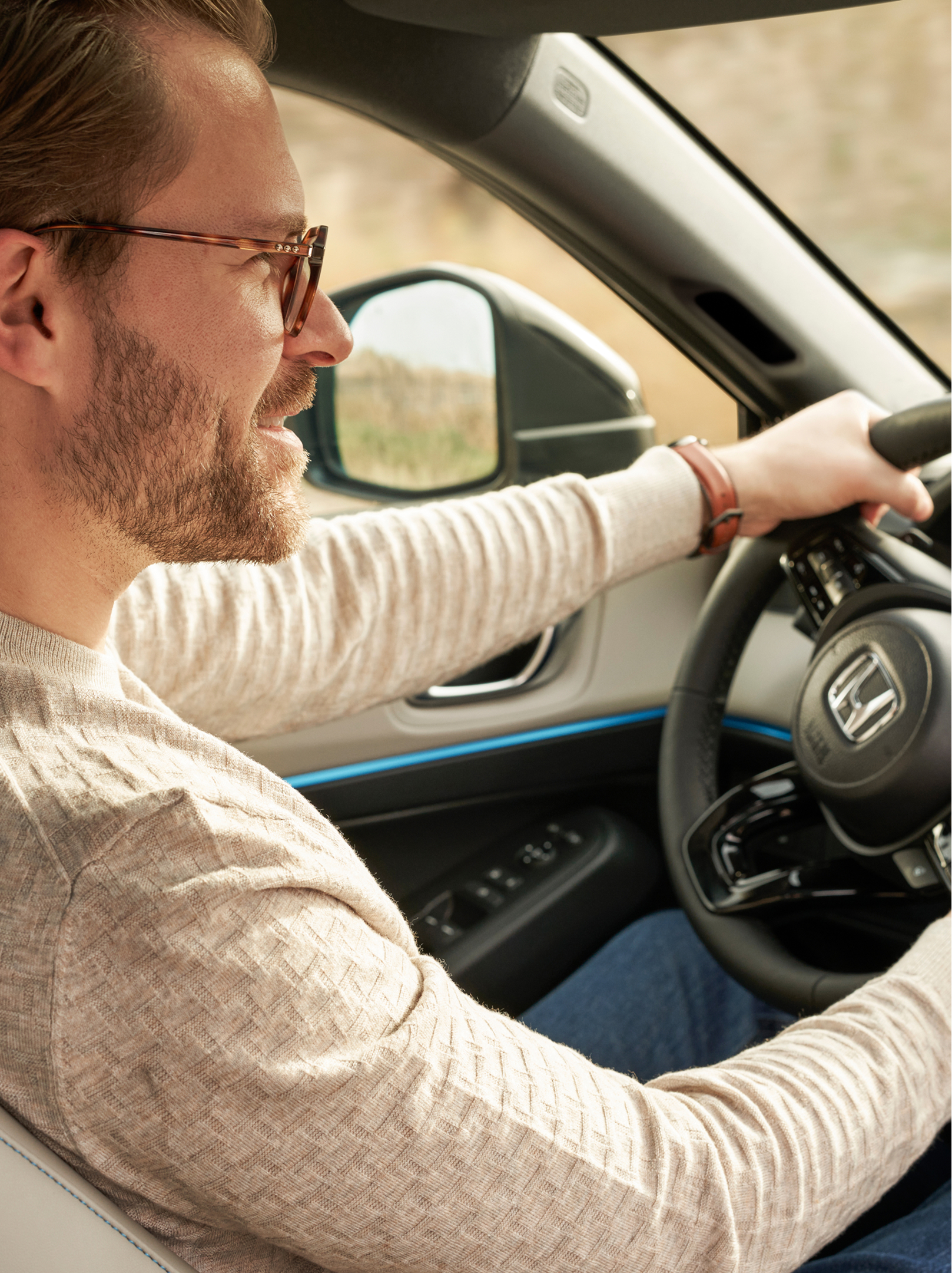 Side view of a person driving a Honda vehicle, visible through the driver's side window. The interior shows part of the Honda steering wheel and dashboard with blue accent lighting.