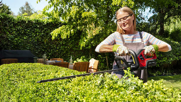 Kvinna som klipper en häck med Hondas batteridrivna häcksax i trädgårdsmiljö.