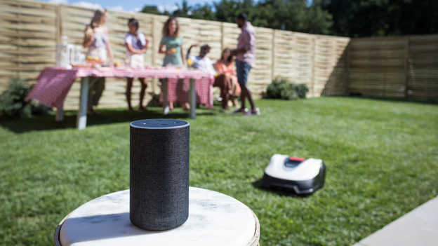 Närbild på Amazon Alexa med Miimo och trädgårdsparty i bakgrunden på gräsmattan.