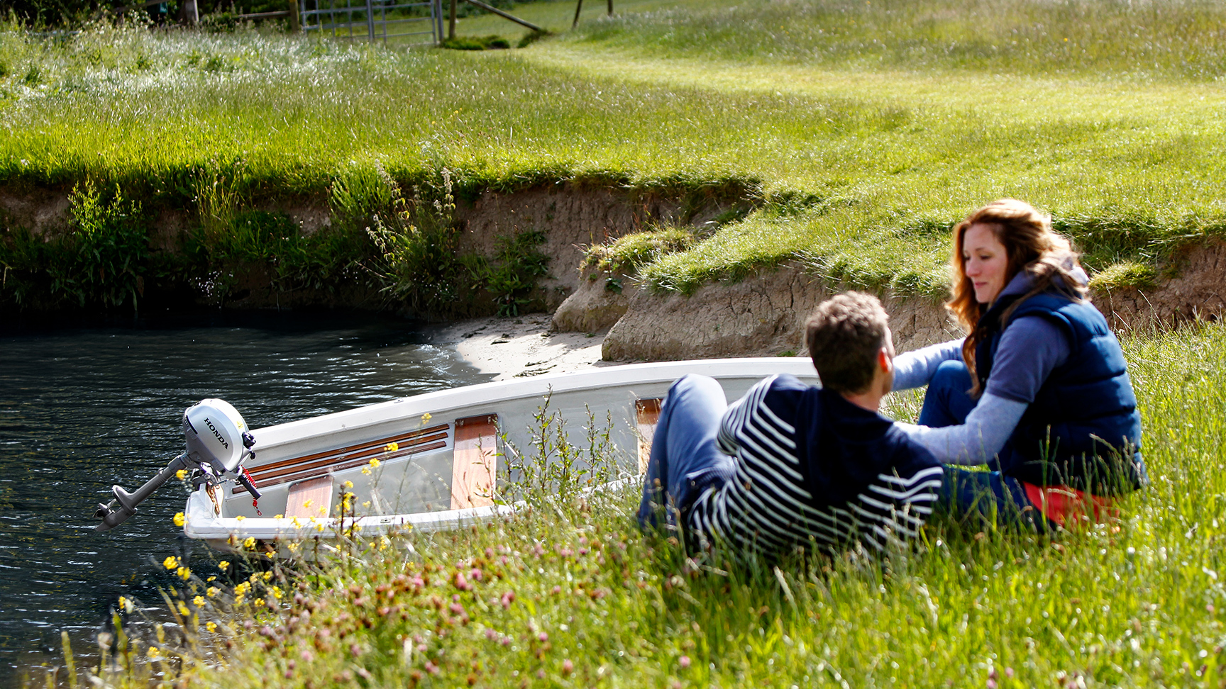 Två människor som sitter på en flodstrand bredvid en båt i vattnet.