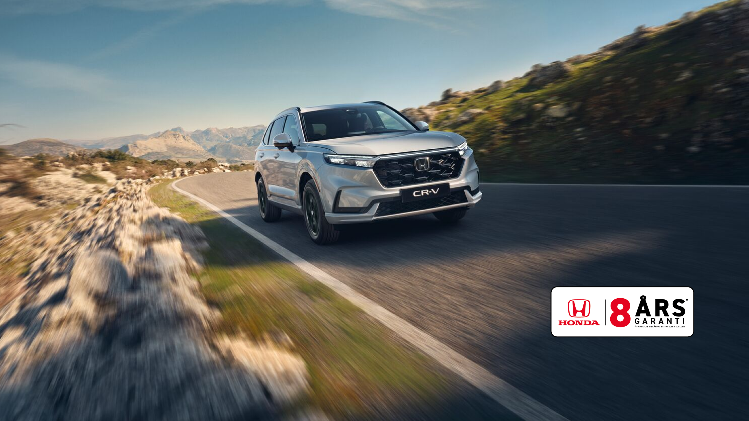 En silvrig Honda CR-V SUV kör på en slingrande bergsväg med karg terräng och berg i bakgrunden. Bilen är i rörelse, vilket skapar en känsla av fart och äventyr i det karga landskapet.