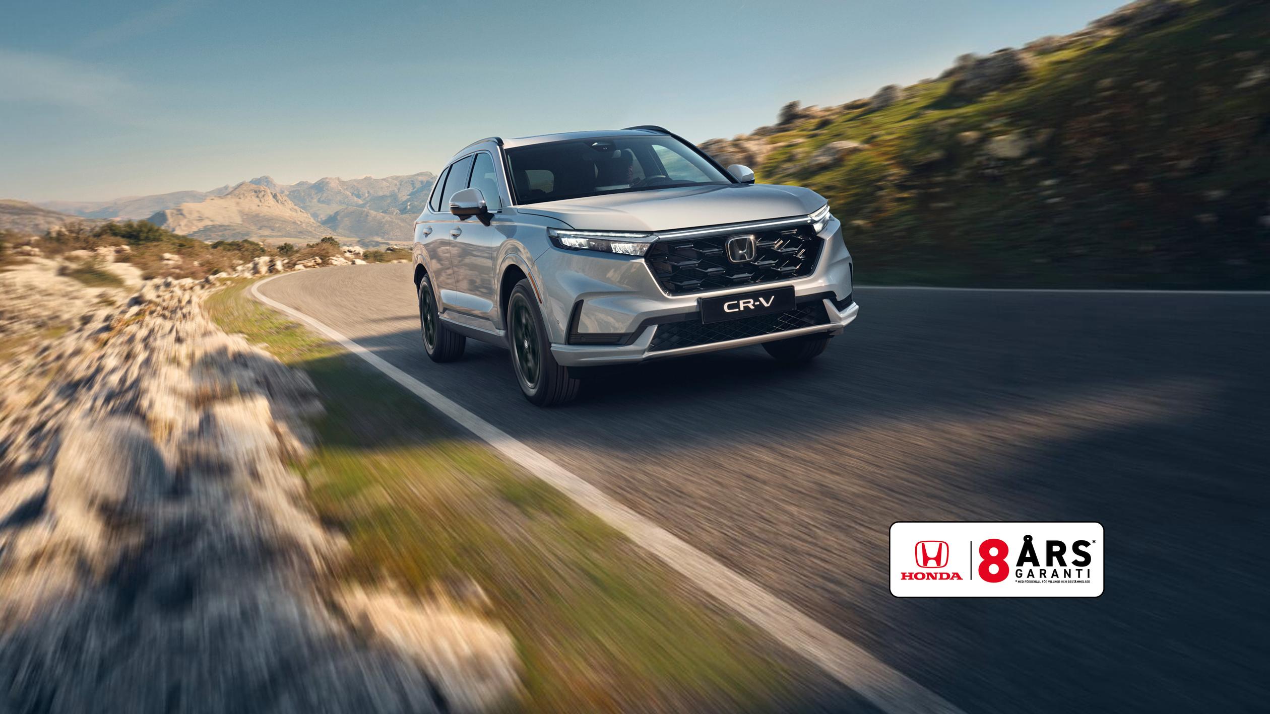 En silvrig Honda CR-V SUV kör på en slingrande bergsväg med karg terräng och berg i bakgrunden. Bilen är i rörelse, vilket skapar en känsla av fart och äventyr i det karga landskapet.