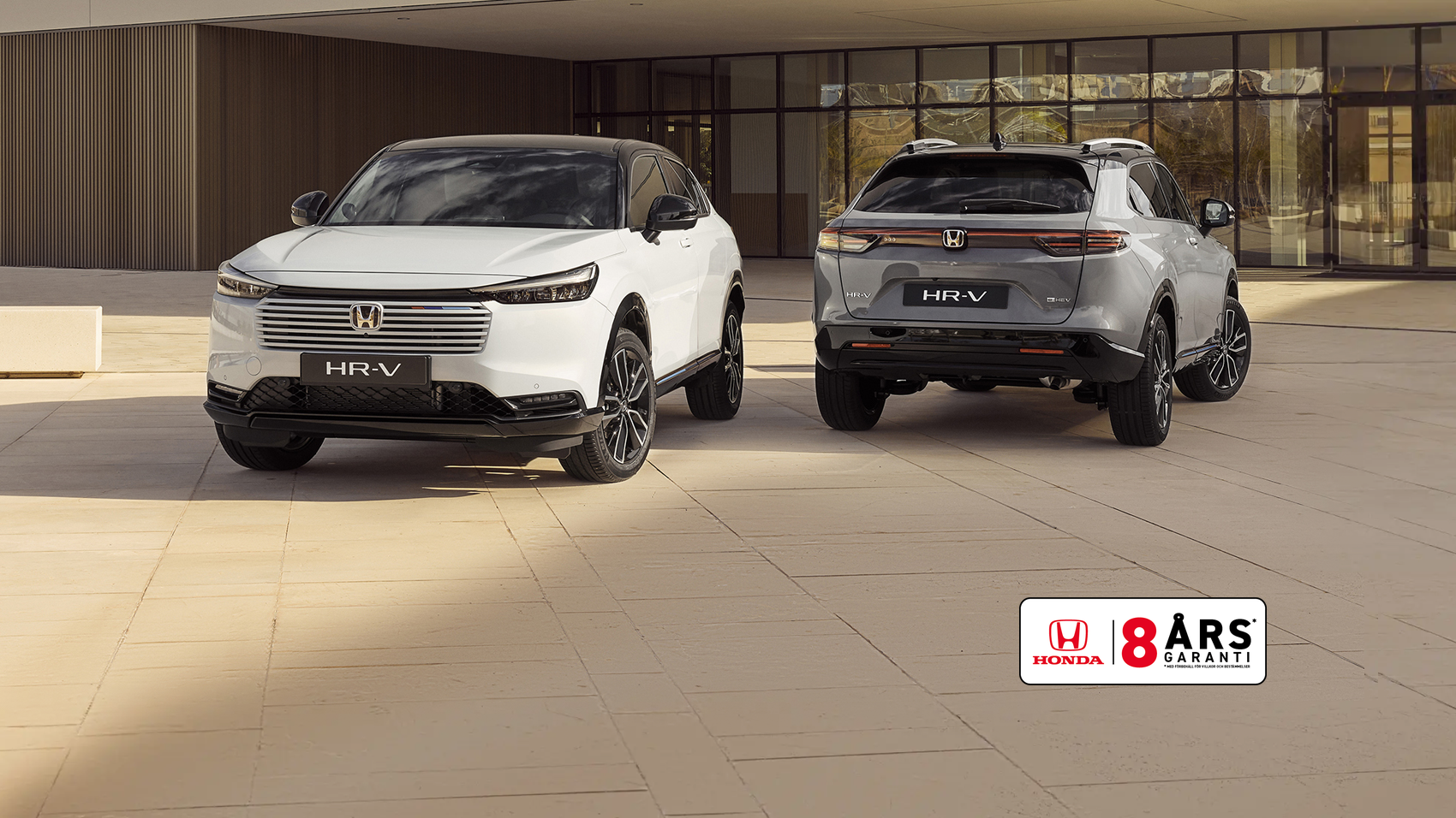 Två Honda HR-V SUV:ar parkerade framför en modern byggnad. Den vita HR-V är i förgrunden vänd framåt, medan den silvriga HR-V är i bakgrunden vänd bortåt, vilket visar bilens fram- och bakdesign.