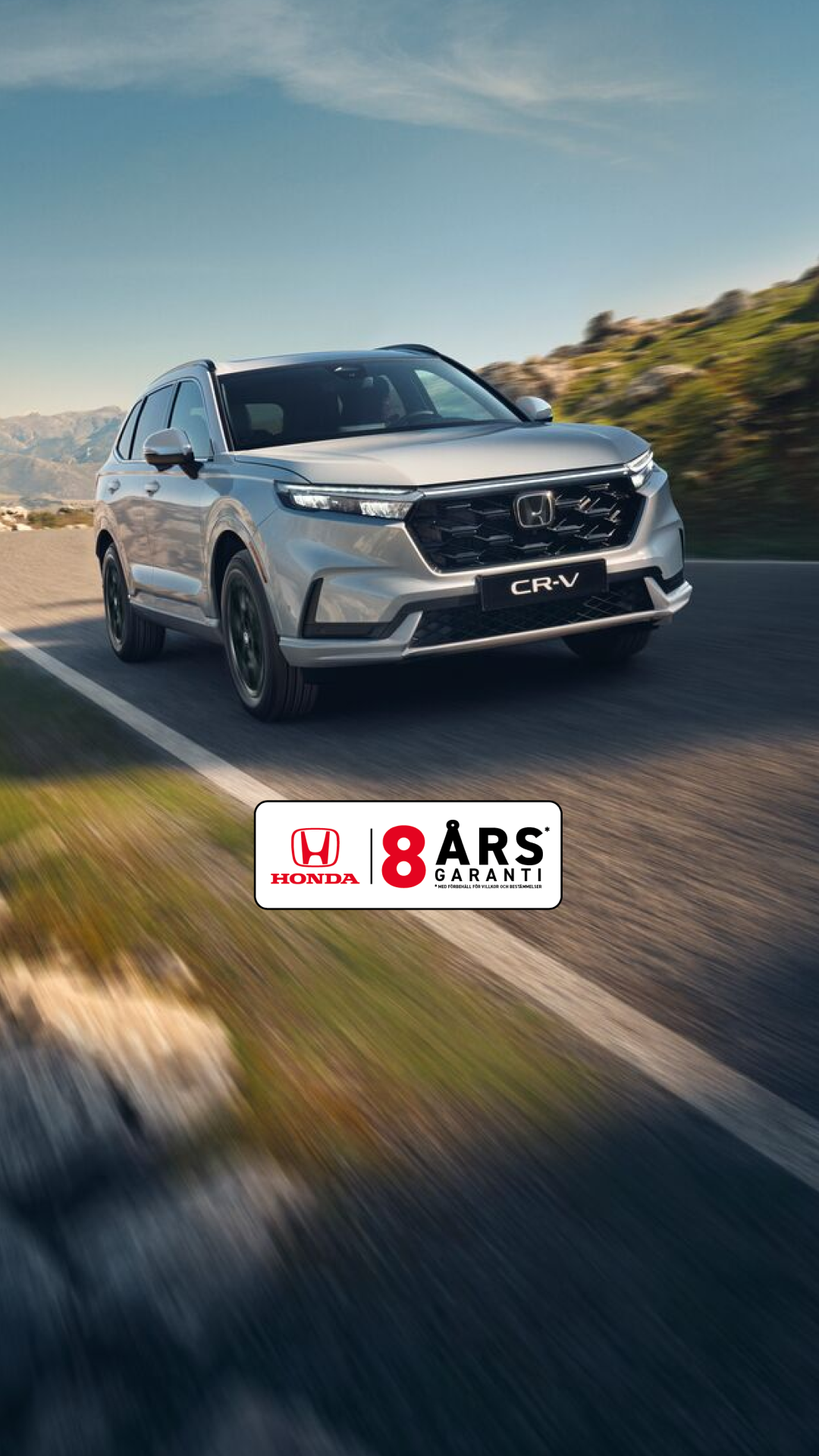 En silvrig Honda CR-V SUV kör på en slingrande bergsväg med karg terräng och berg i bakgrunden. Bilen är i rörelse, vilket skapar en känsla av fart och äventyr i det karga landskapet.