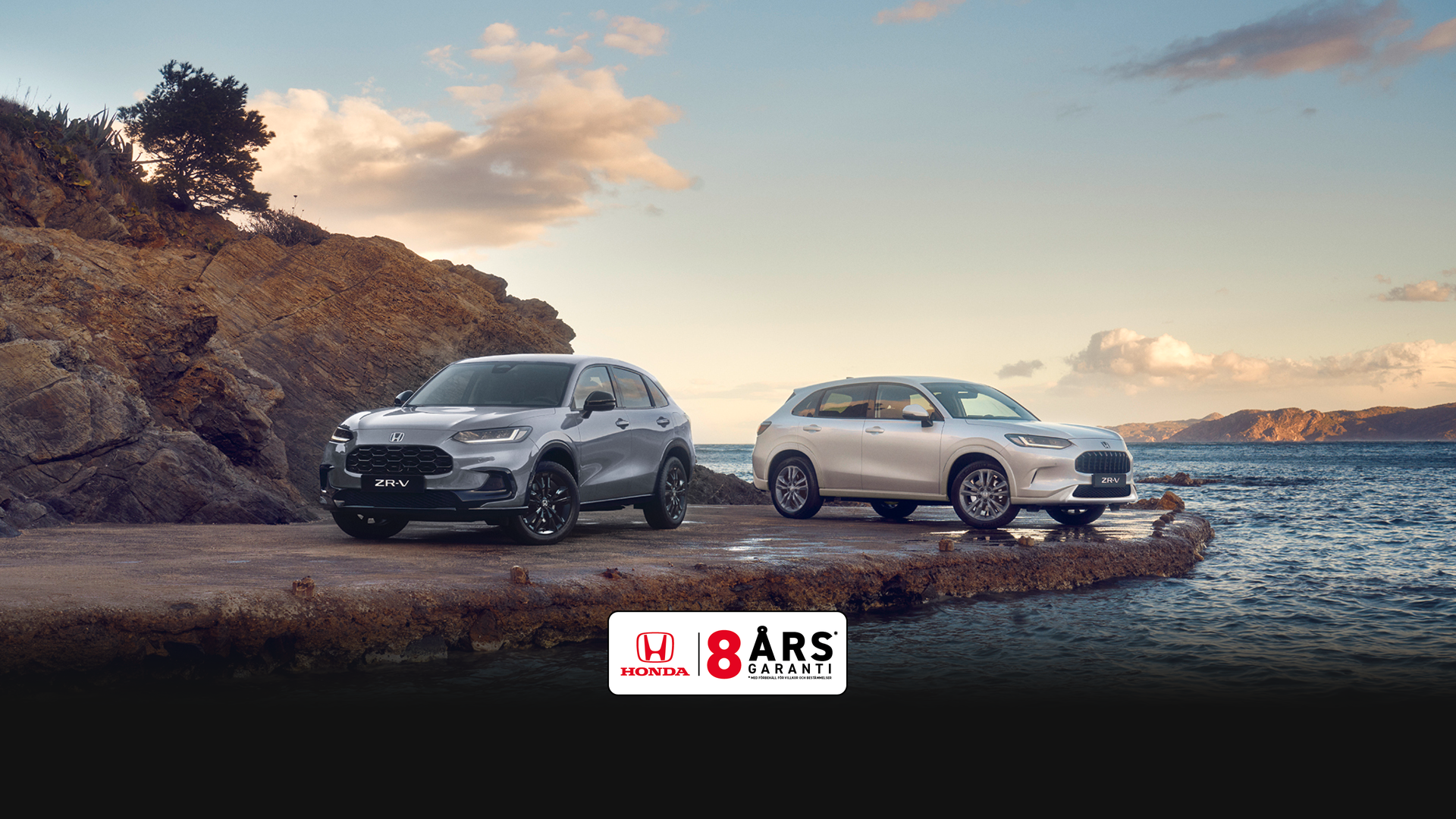 Two Honda ZR-V SUVs, one blue and one white, parked on a rocky coastal outcrop with water and hills in the background under a partly cloudy sky.